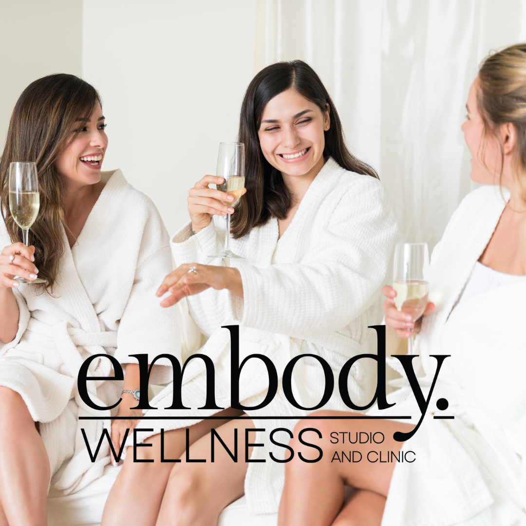 3 women sitting in a spa, the room is bright and cheery. All 3 women have a glass of champagne and the middle woman is showing off an engagement ring.